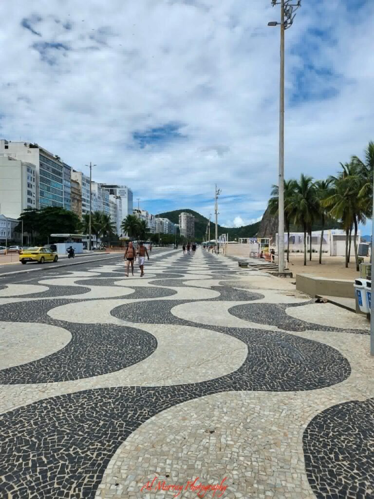 The black and white wave patterned promenade stretches along Copacabana Beach, lined with palm trees, sandy shoreline, and city buildings beneath a bright cloudy sky. This iconic waterfront scene showcases one of the memorable Atlantic crossing cruise ports where travelers can step ashore and explore Rio’s vibrant coastline