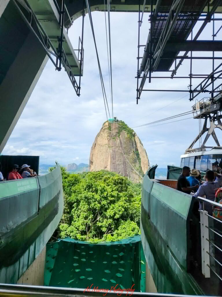 Cable cars wait at the station as visitors board for a ride toward Sugarloaf Mountain, with lush green forest and sweeping city views in the distance. The dramatic perspective and iconic Rio landscape highlight the kind of unforgettable shore excursion that often stands out in a transatlantic cruise review.