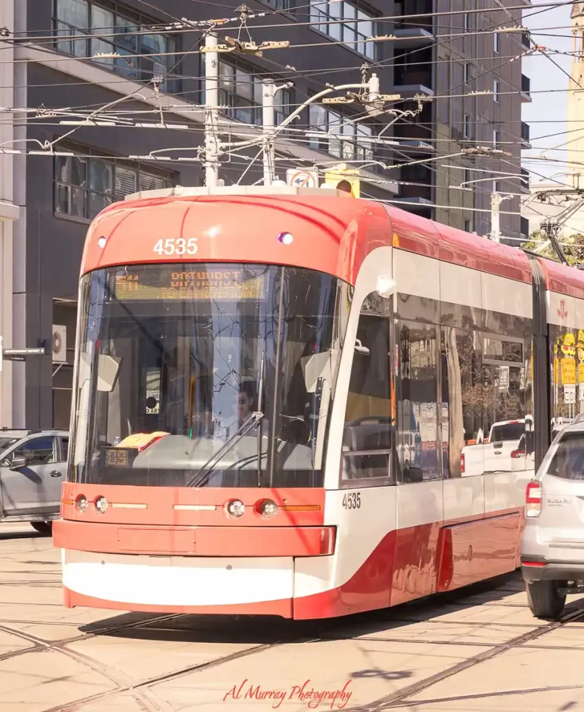Red and white Toronto streetcar traveling through a downtown intersection surrounded by condo buildings and overhead transit wires on a sunny day. The busy city setting captures the experience of a self guided Toronto food tour using the streetcar to explore restaurants and neighbourhoods across the city.