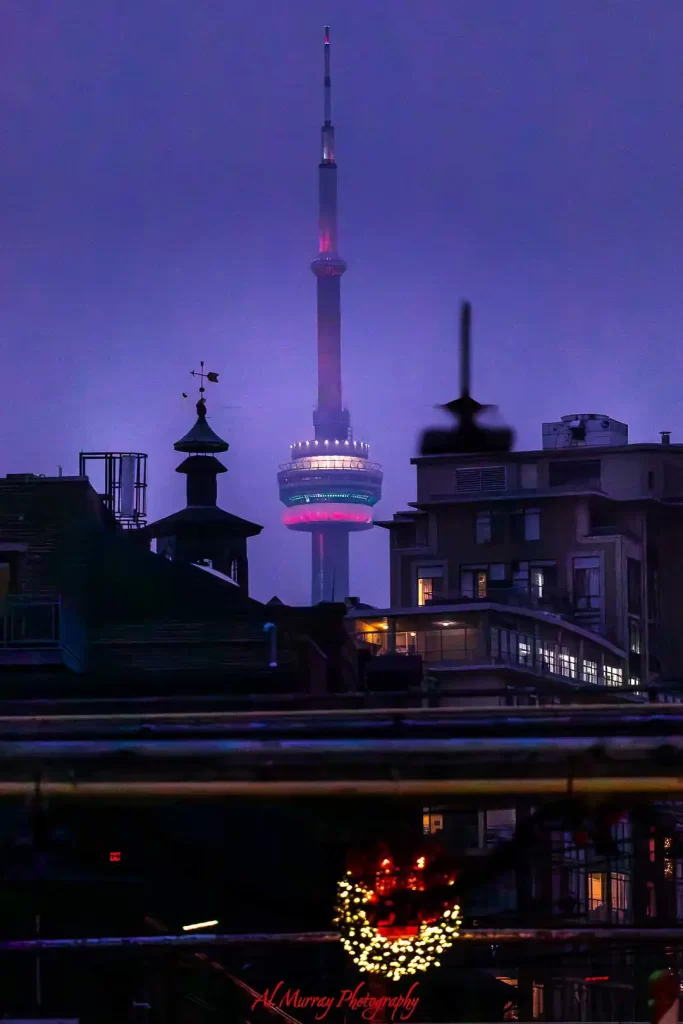 CN Tower glowing through purple evening fog above downtown Toronto buildings with warm city lights visible below. The moody skyline view captures the atmosphere of a Toronto food tour by streetcar after dark as visitors explore the city between neighbourhood dining stops.