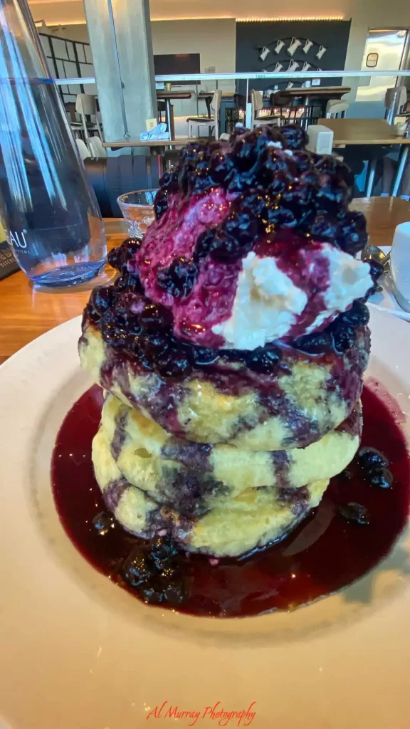 Tall stack of fluffy blueberry pancakes covered in warm blueberry sauce, whipped cream, and whole blueberries on a white plate inside a modern restaurant. The rich brunch dish highlights a stop along the best Toronto streetcar route for food lovers exploring cafes and local dining spots across the city.