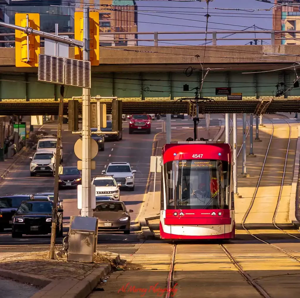 Red Toronto streetcar traveling along tracks beneath an overpass with downtown traffic moving beside it in warm evening light. Overhead transit wires and city streets create an urban scene that reflects the experience of a Toronto food tour by streetcar.