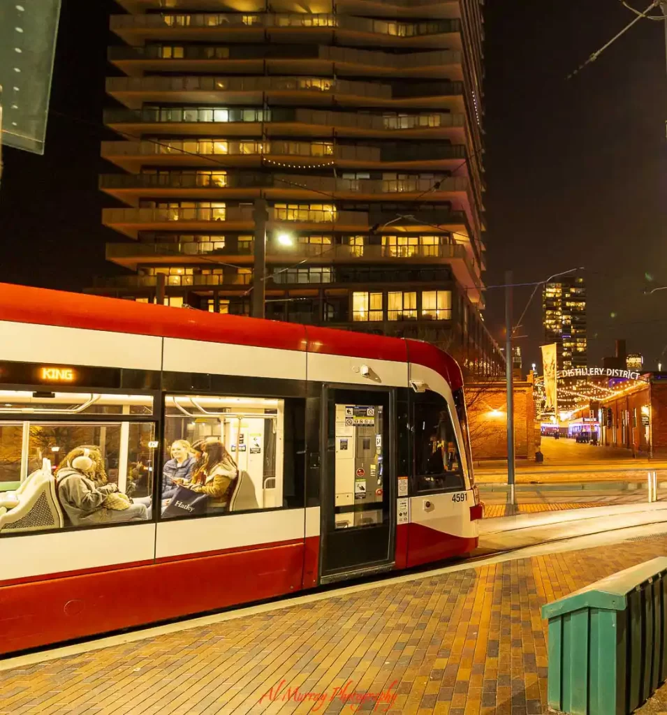 Red and white Toronto streetcar stopped at night near the Distillery District with illuminated condo buildings and glowing streetlights in the background. Passengers sit inside the streetcar as it travels through one of Toronto food neighbourhoods known for restaurants, bars, and evening city life.