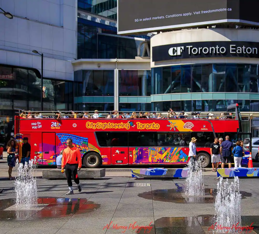Bright red double decker sightseeing bus driving past CF Toronto Eaton Centre on a sunny day with people walking through the downtown plaza and fountains spraying in the foreground. The busy city scene highlights the energy of hop-on hop-off bus tours exploring Toronto landmarks and attractions.
