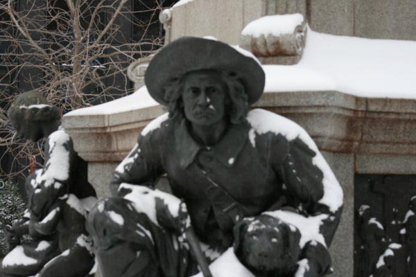 Snow covers a bronze statue of a seated man wearing a wide brimmed hat with a second figure behind him and a stone monument in the background. The wintry public artwork creates a striking outdoor stop for Canada fall and winter getaways.