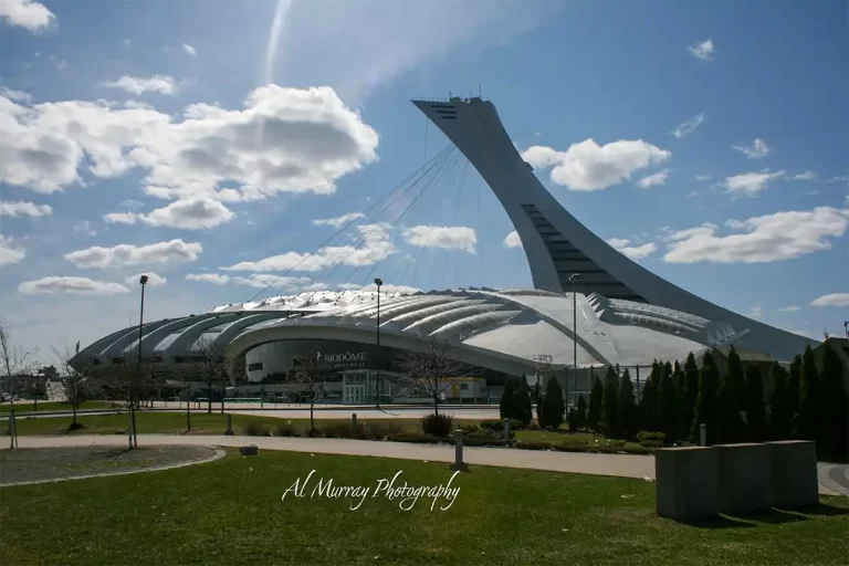 Montreal Olympic Stadium and its leaning observation tower rise behind the silver roof of the Biodome beneath a bright blue sky with scattered clouds. The landmark highlights a Montreal stop on a Montreal Quebec City and Ottawa itinerary and includes the visible text "BIODÔME DE MONTRÉAL" and "Al Murray Photography".