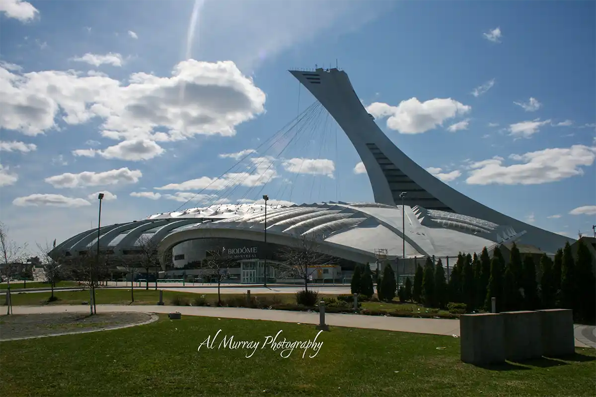 Montreal Olympic Stadium and its leaning observation tower rise behind the silver roof of the Biodome beneath a bright blue sky with scattered clouds. The landmark highlights a Montreal stop on a Montreal Quebec City and Ottawa itinerary and includes the visible text "BIODÔME DE MONTRÉAL" and "Al Murray Photography".
