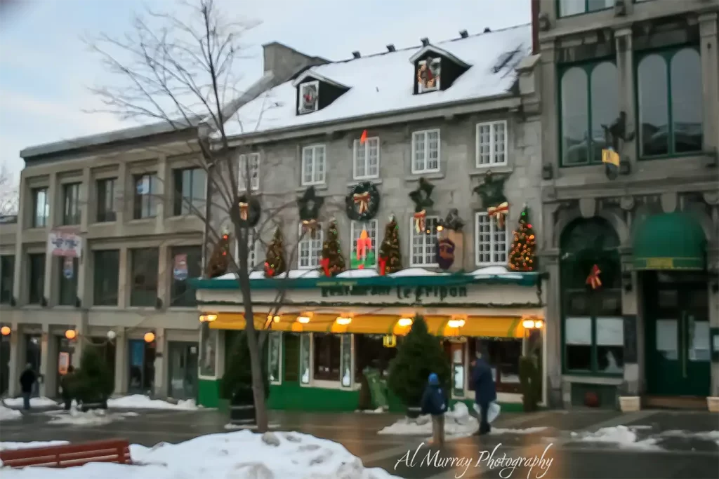 A snow covered historic stone restaurant glows with yellow awnings and Christmas wreaths as pedestrians walk along the wet winter sidewalk. The festive Montreal street scene helps answer "Is winter a good time to visit Canada" and visible text includes "Jardin Nelson" and "Al Murray Photography".