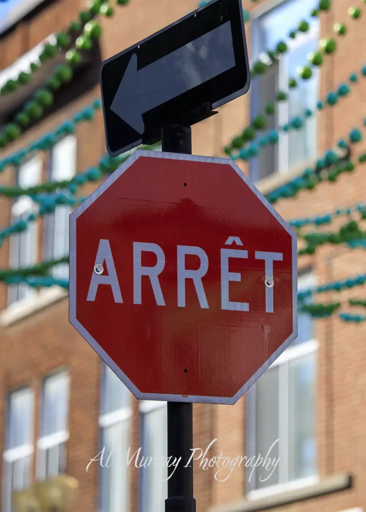 A red stop sign marked "ARRÊT" stands below a black left arrow sign in front of a brick building decorated with green garlands. The French road sign reflects the language visitors may encounter when asking "do I need to know French to visit Montreal Quebec" and the watermark reads "Al Murray Photography".
