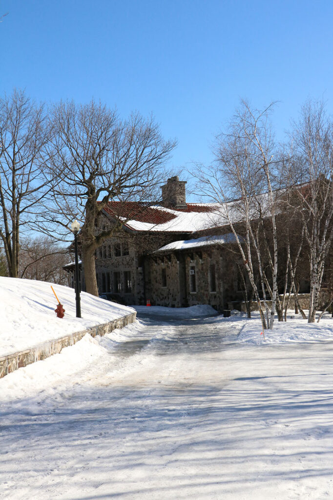 A rustic stone lodge with a red roof sits among bare trees beside a snow covered path under a clear blue sky. The peaceful setting reflects the cozy scenery found in some of the best winter destinations in Canada.