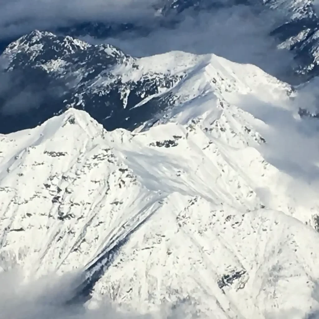 Snow covered mountain peaks rise through low gray clouds with dark rocky ridges visible between bright white slopes. The dramatic alpine landscape highlights the scenery available on Canadian winter trips for non skiers.