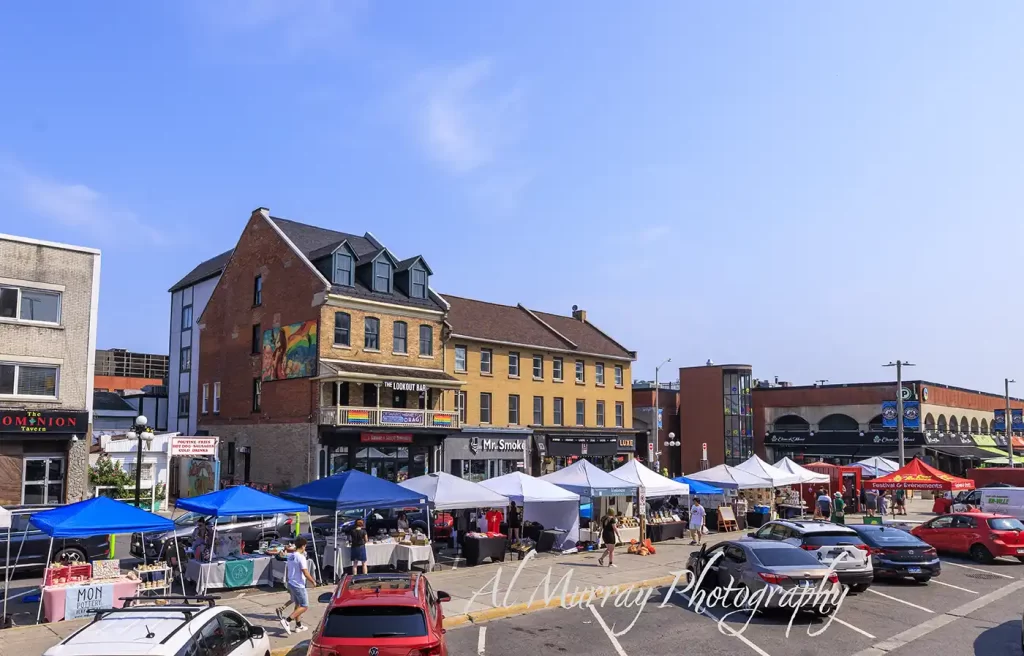 Outdoor vendor tents fill a sunny street at Ottawa ByWard Market as shoppers browse handmade goods beside parked cars and historic brick storefronts. Visible text includes "The Dominion Tavern" "The Lookout Bar" "Ottawas longest running drag bar" "Mr Smoke" "LUXY" "Poutine Fries Hot Dogs Sausages Cold Drinks" "Festival and Evenements" "Mon Pottery" and "Al Murray Photography"