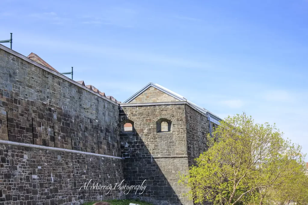 Historic stone fortification walls and an adjoining building rise beneath a bright blue sky with fresh green trees along the base. The preserved architecture highlights a Quebec City stop on a Montreal Quebec City and Ottawa itinerary and the watermark reads "Al Murray Photography".
