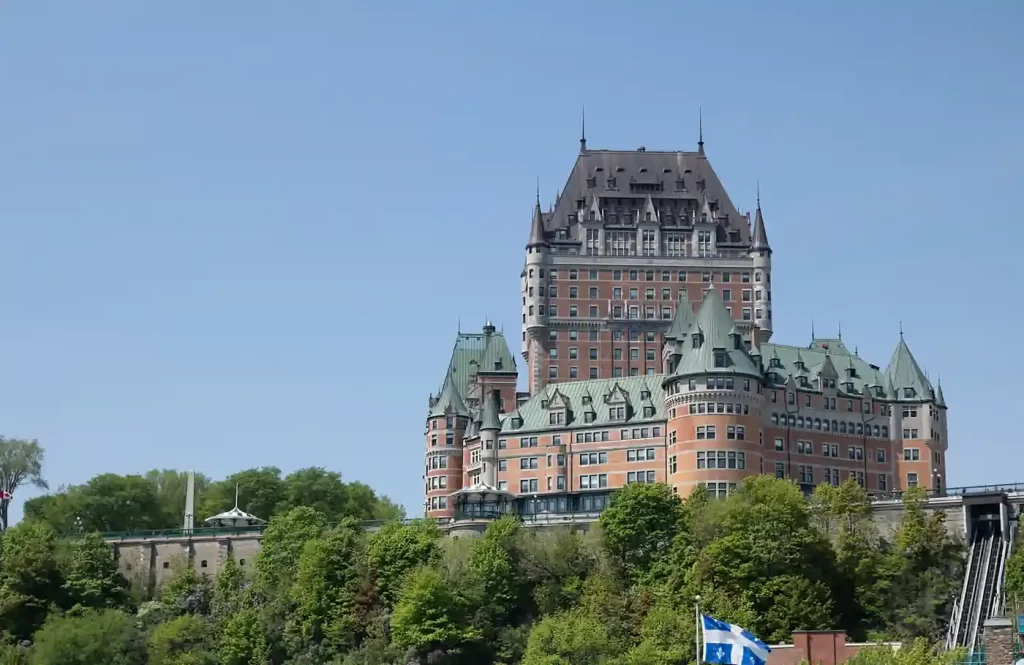 Fairmont Le Château Frontenac rises above green trees and historic stone walls in Quebec City beneath a clear blue sky. Its copper roofs and castle like towers highlight a landmark often featured among the best places to visit in Canada in fall and winter.