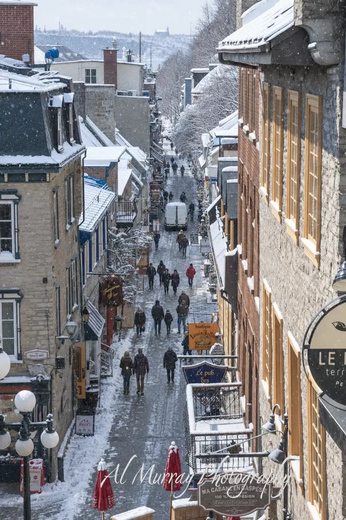 A narrow pedestrian street in Old Quebec slopes between snow covered stone and brick buildings as people walk past shops and restaurants during falling snow. The wintry Quebec City scene is a scenic stop on a Montreal Quebec City and Ottawa itinerary. Visible text includes "Sous le fort" "Fumeron d Or" "boutique Louis Jolliet souvenirs" "Le pub des" "biz" "evian" "100 pour cent canadien produit véritable" and "Al Murray Photography"