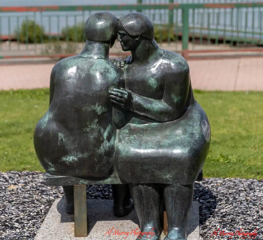 A dark bronze sculpture shows two seated figures leaning closely together with their foreheads touching during a Windsor Essex fall getaway. The artwork sits on a gravel base beside green grass and includes the red watermark "Al Murray Photography".