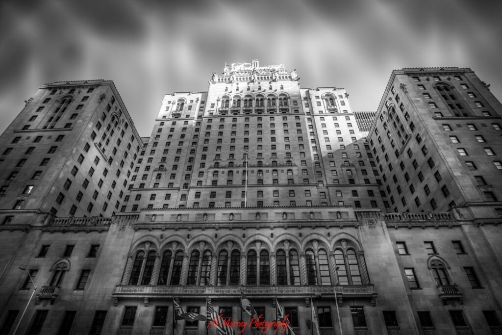Black and white low angle view of the historic Fairmont Royal York hotel in downtown Toronto with its tall stone facade and rows of arched windows rising beneath a cloudy sky. The landmark offers a traditional downtown accommodation setting for travellers deciding between a hotel or vacation rental in Toronto.