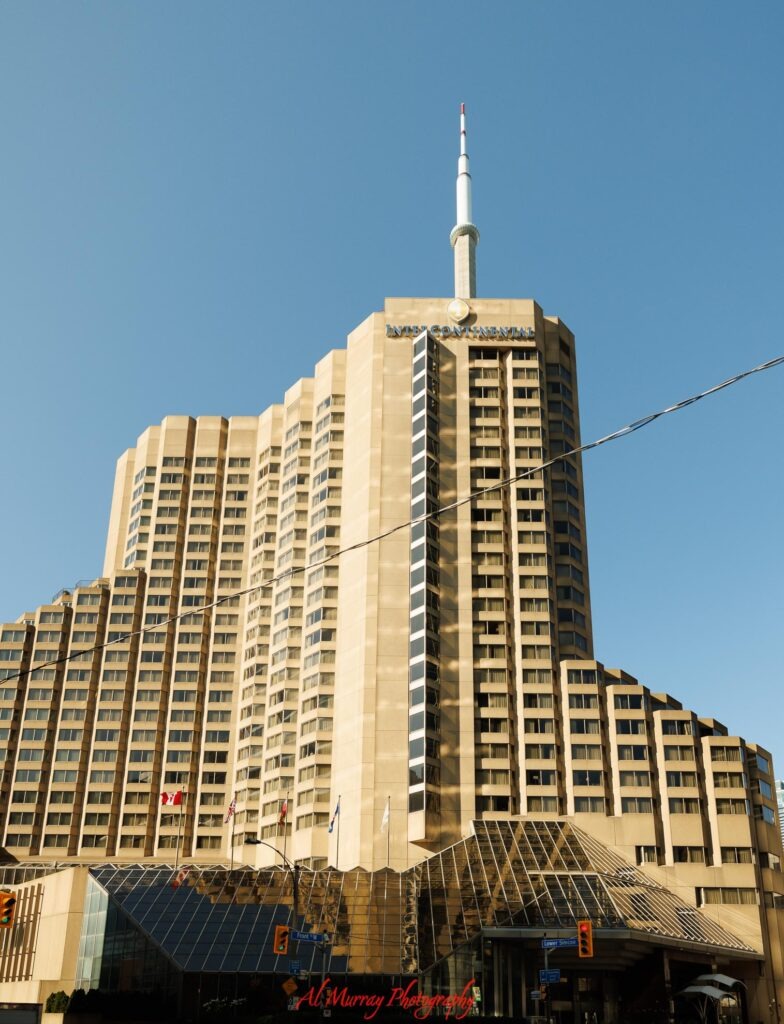 Tall InterContinental hotel building rising against a clear blue sky with a glass entrance canopy and Canadian flags at street level. Visible signs read "INTERCONTINENTAL" "Front W" and "Lower Simcoe" giving travellers a downtown hotel example when comparing hotel vs VRBO Toronto.