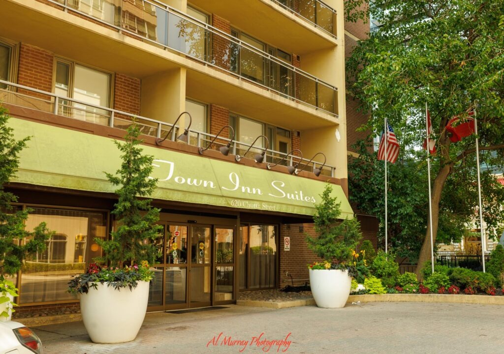 Exterior of Town Inn Suites in Toronto with balconies. landscaped planters. and flags beside the entrance. The awning reads "Town Inn Suites" and "620 Church Street". making the hotel relevant for travellers looking for Toronto hotels with kitchenettes.