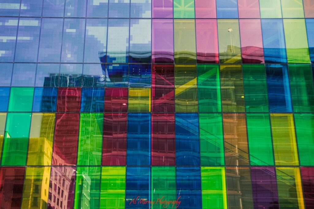 Colourful glass panels in shades of blue green yellow and pink cover a modern building in Montreal and reflect the surrounding city architecture. The vibrant downtown streetscape offers a sense of the urban setting for travellers deciding where to stay in Montreal for mature travellers