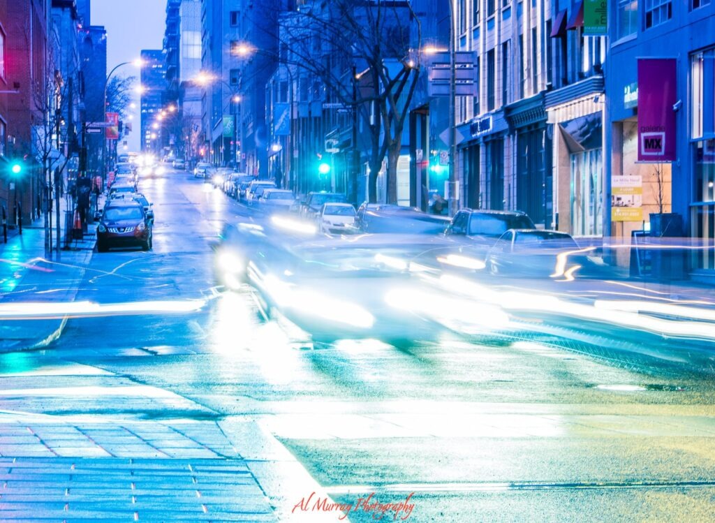 A busy Montreal street at dusk with glowing traffic lights and long exposure car light trails stretching between tall buildings. Signs reading "galerie MX" and "Le Mille Neuf" add to the lively downtown setting for travellers considering the best neighbourhood to stay in Montreal.