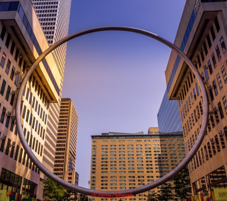 The giant metal Ring sculpture at Esplanade PVM frames office buildings and a clear blue sky in downtown Montreal. The landmark highlights the modern city center for travelers comparing the best neighbourhood to stay in Montreal.