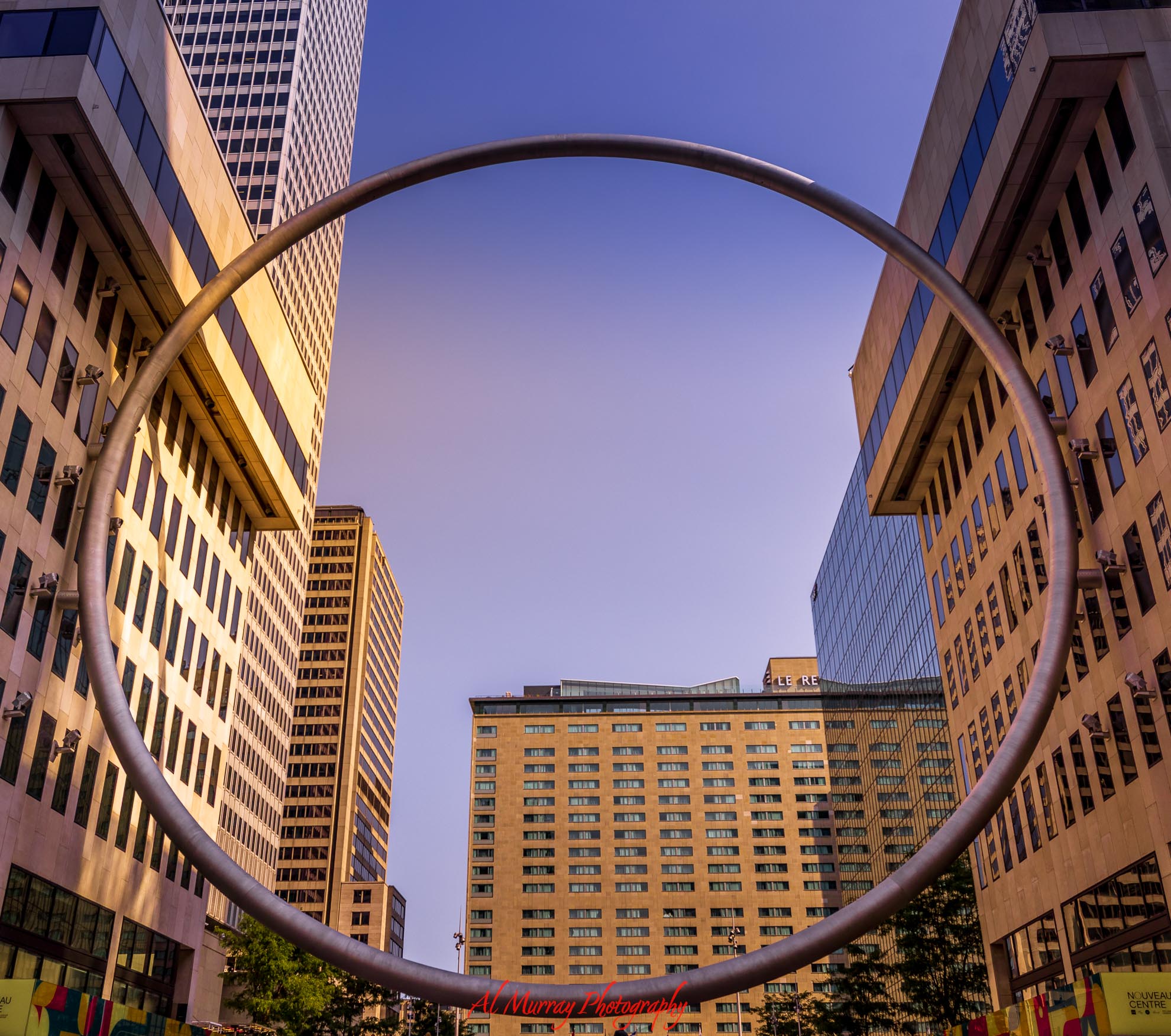 The giant metal Ring sculpture at Esplanade PVM frames office buildings and a clear blue sky in downtown Montreal. The landmark highlights the modern city center for travelers comparing the best neighbourhood to stay in Montreal.