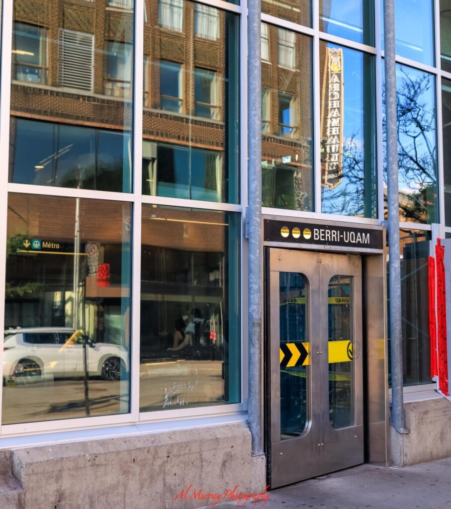 Entrance to the Berri UQAM metro station in Montreal with glass windows reflecting nearby brick buildings and a street scene. Signs read "BERRI UQAM" and "Métro" showing the convenient transit access travelers may consider when comparing the best neighbourhoods to stay in Montreal.