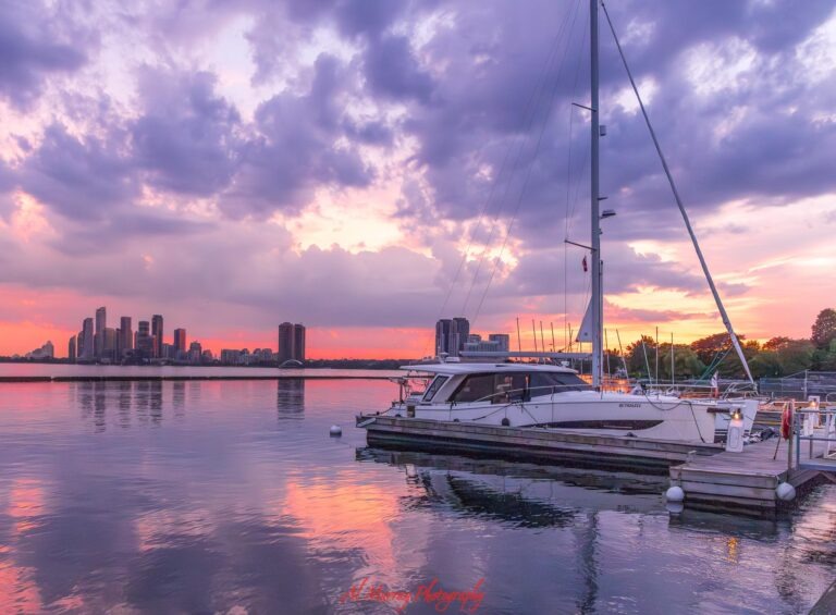 Sailboat docked at a waterfront marina during a pink and purple sunset with a city skyline across the calm water. The peaceful waterfront setting provides travel context for readers comparing Hotel vs VRBO Toronto.