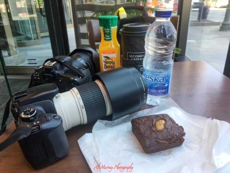 A travel breakfast of a chocolate brownie coffee Simply Orange juice and Eska water sits on a wooden table beside two DSLR cameras with large lenses. Readable labels include "Simply Orange" "SECOND CUP" "eska" and the watermark "Al Murray Photography". The breakfast and camera gear add a personal travel moment to a guide about Toronto hotels with breakfast included.