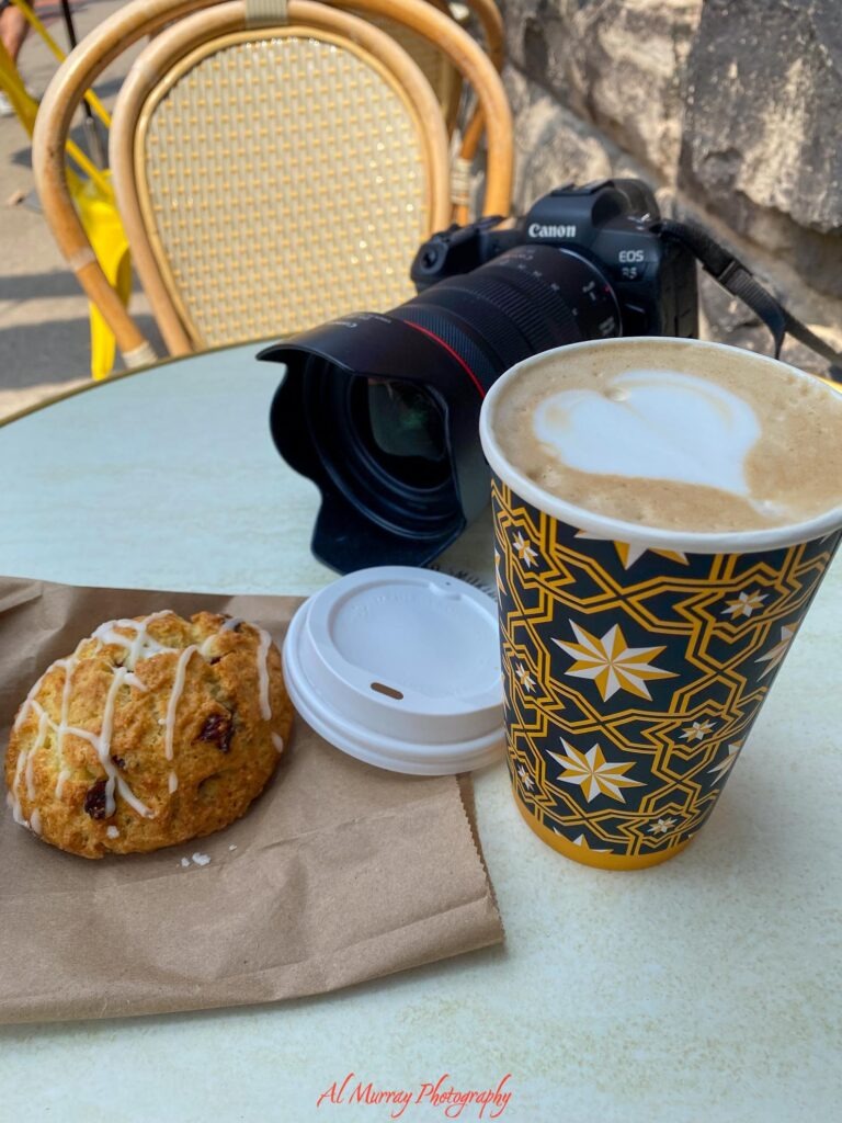 toronto hotels with breakfast included A patterned takeaway cup of latte with heart shaped foam art and an iced cranberry scone sit on an outdoor cafe table beside a Canon EOS R5 camera. The casual breakfast stop complements a travel guide about downtown Toronto hotels with breakfast included. Visible text reads "Canon" "EOS R5" and "Al Murray Photography".