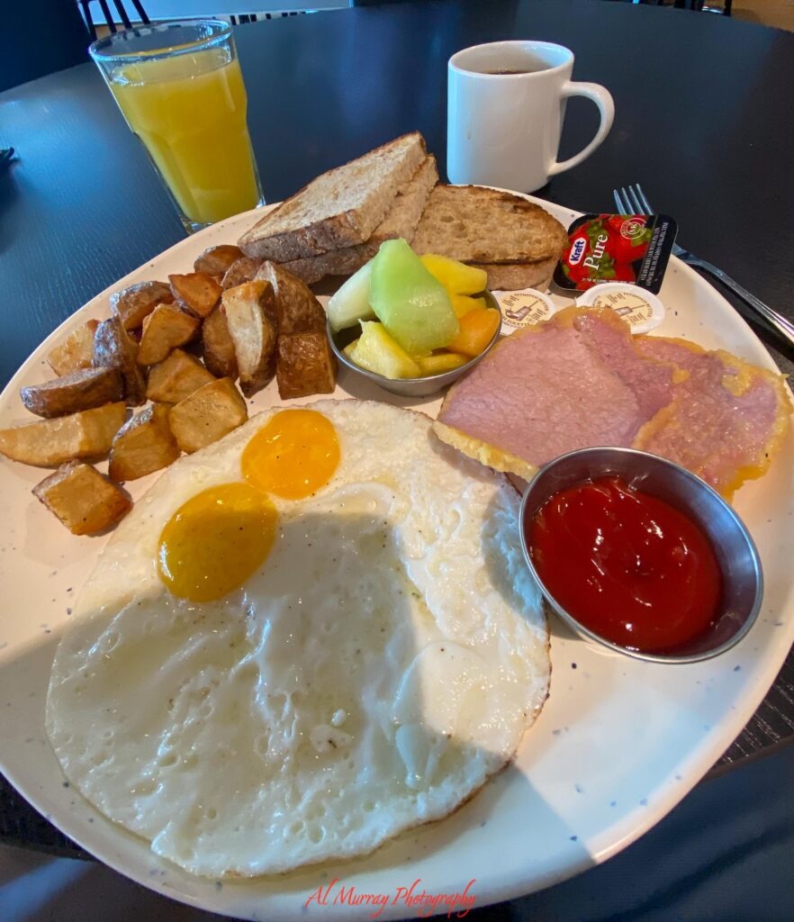 toronto hotels with breakfast included A full hotel breakfast plate with two fried eggs ham roasted potatoes toast fresh melon ketchup and jam sits beside a glass of orange juice and a mug of coffee. The generous meal highlights the breakfast travelers can expect when researching Toronto hotels with breakfast included. Visible text reads "Kraft Pure" and "Al Murray Photography".