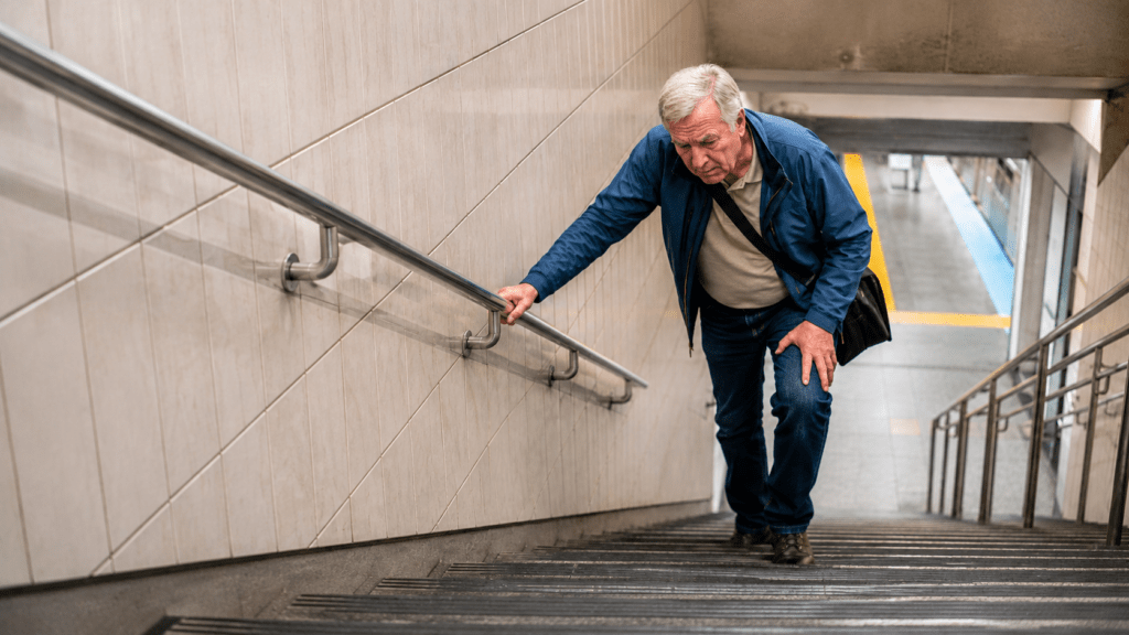 An older man climbs a long transit station staircase while gripping the handrail and resting one hand on his knee. The steep steps highlight an important accessibility concern when deciding where to stay in Toronto with limited mobility.