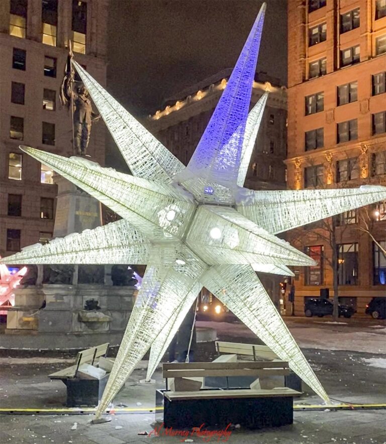 A large illuminated white star sculpture glows at night in a snowy downtown square surrounded by tall historic buildings and benches. The winter streetscape highlights the kind of central location and seasonal setting to consider in a Montreal hotel booking checklist. "Al Murray Photography".