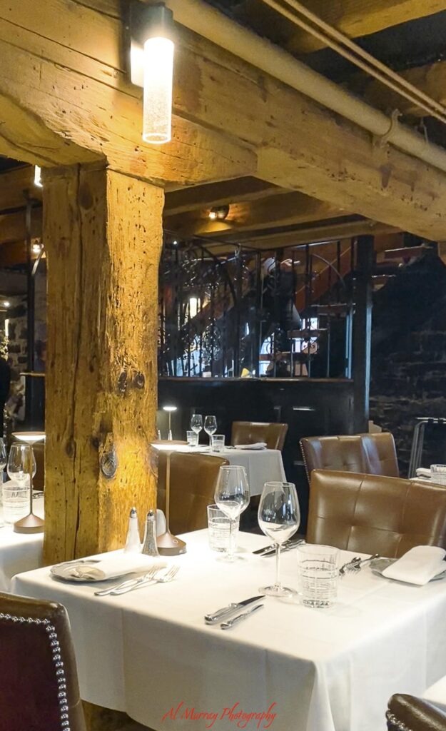 Warm restaurant interior with exposed wooden beams stone walls brown leather chairs and white tablecloths set with wine glasses. Nearby dining options like this can be a useful consideration when choosing a Montreal short term rental. "Al Murray Photography".