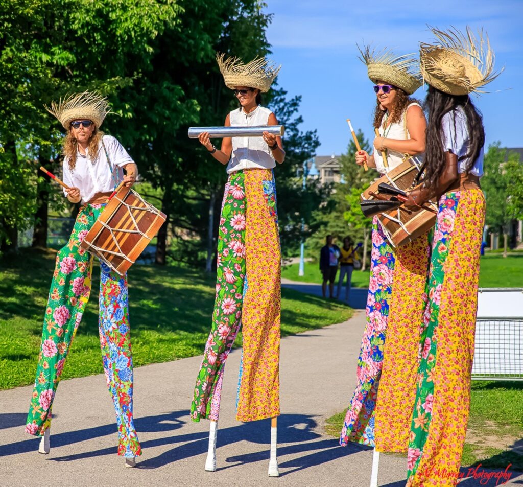where to stay in Toronto as a solo traveller Four street performers on tall stilts wear wide straw hats and bright floral pants while playing drums and metal percussion outdoors.