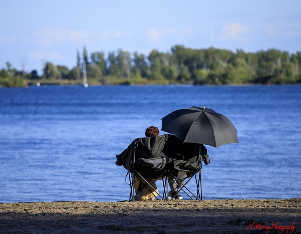 where to stay in Toronto as a solo traveller A person relaxes in a folding chair beneath a black umbrella on a sandy Toronto waterfront with calm blue water and trees in the distance.