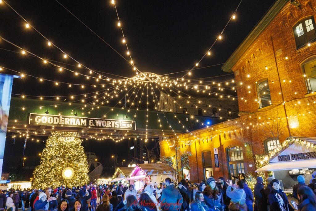 A crowded holiday market glows beneath strings of warm lights and a large illuminated Christmas tree outside red brick buildings at the Distillery District in Toronto. Signs read “GOODERHAM & WORTS LIMITED” and “MINI PANCAKES” while red script at the bottom reads “Al Murray Photography”.