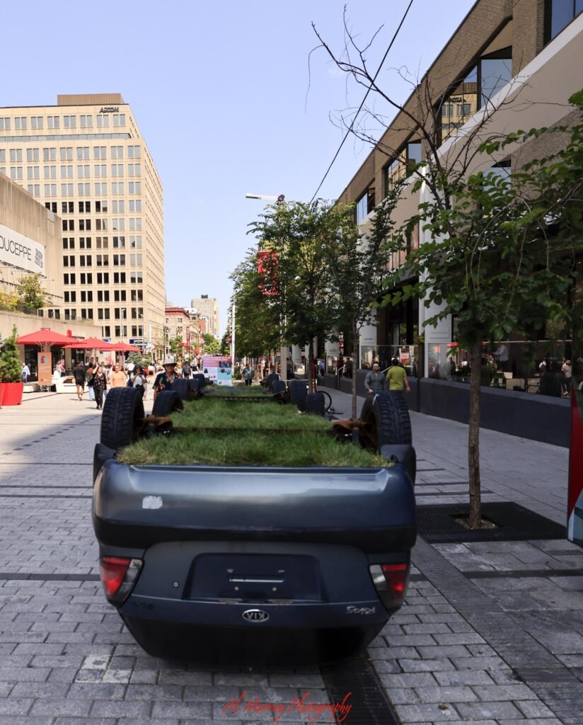 how much do hotels cost in Montreal A downtown Montreal pedestrian street is lined with shops patios trees and people walking beneath a clear blue sky. An upside down car filled with grass serves as a quirky planter in the foreground and adds local context for travelers researching how much do hotels cost per night in Montreal.