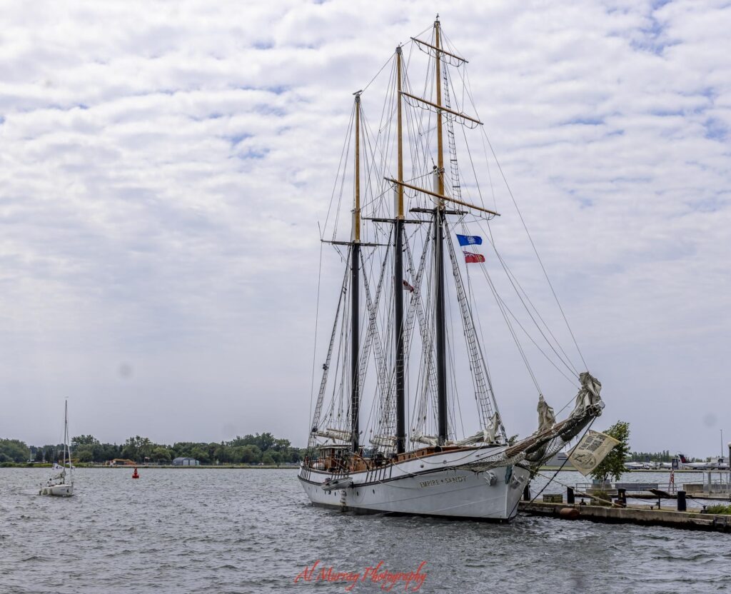 The tall masted Empire Sandy is docked along the Toronto waterfront while a small sailboat crosses the harbor under a cloudy sky. The harbor scene gives travelers deciding where to stay in Toronto for couples a look at the waterfront atmosphere. Text on the ship reads “EMPIRE SANDY” and red script at the bottom reads “Al Murray Photography”.