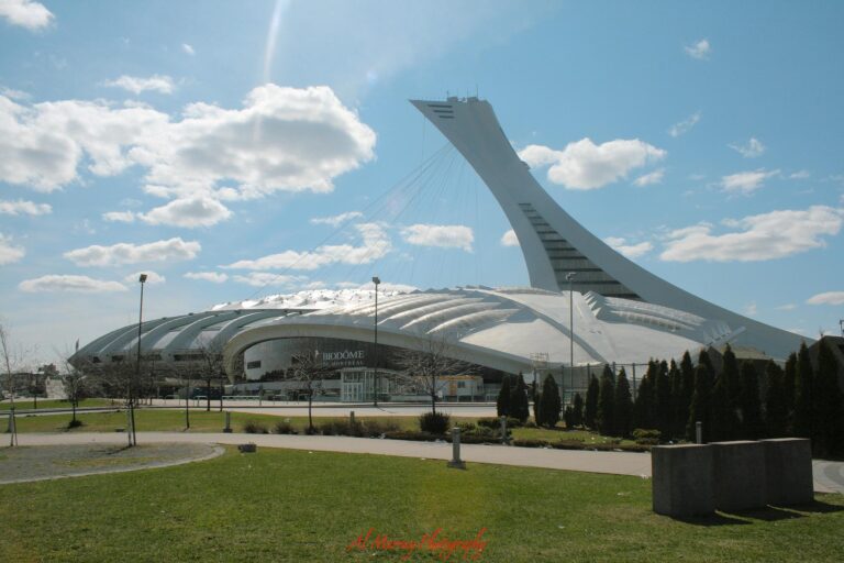 The Montreal Biodome and Montreal Olympic Stadium tower under a bright blue sky with scattered clouds. This major attraction in the Olympic Park area highlights a different side of the city when comparing Old Montreal vs Downtown Montreal.
