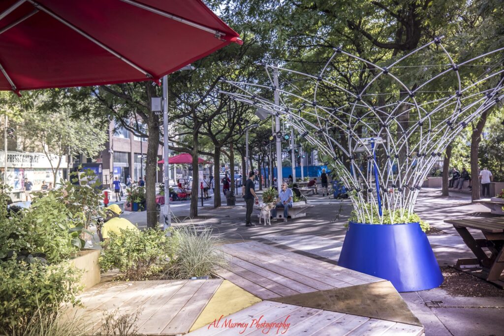 Shaded pedestrian space in Downtown Montreal with leafy trees. red umbrellas. wooden seating. planters. and a large metal public art installation. People relax and walk through this lively urban gathering space between Old Montreal and Downtown Montreal.