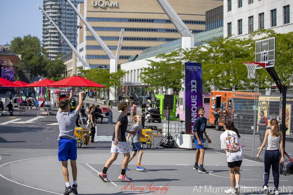 People play basketball on an outdoor court in Downtown Montreal surrounded by food trucks. red umbrellas. trees. and modern buildings. The active public space shows the lively atmosphere visitors can expect when deciding should I stay in Old Montreal or Downtown.