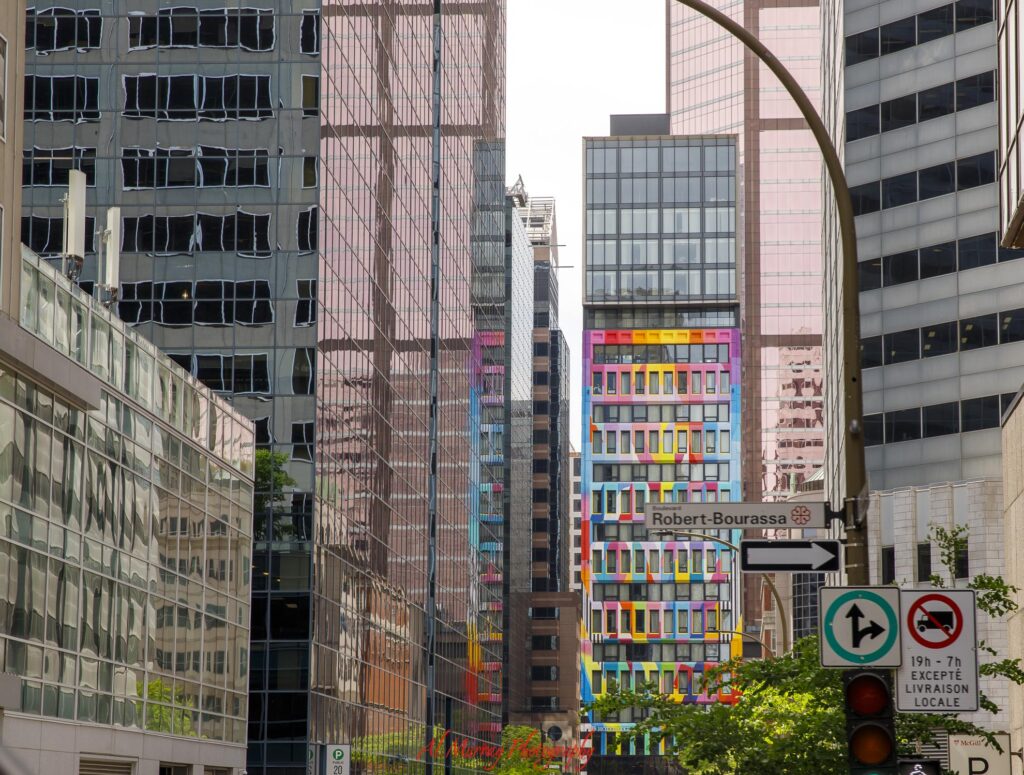 Downtown Montreal skyscrapers line Boulevard Robert Bourassa with reflective glass facades and a brightly painted multicolored building in the distance. The dense modern streetscape provides context for travelers wondering how far is Old Montreal from Downtown.