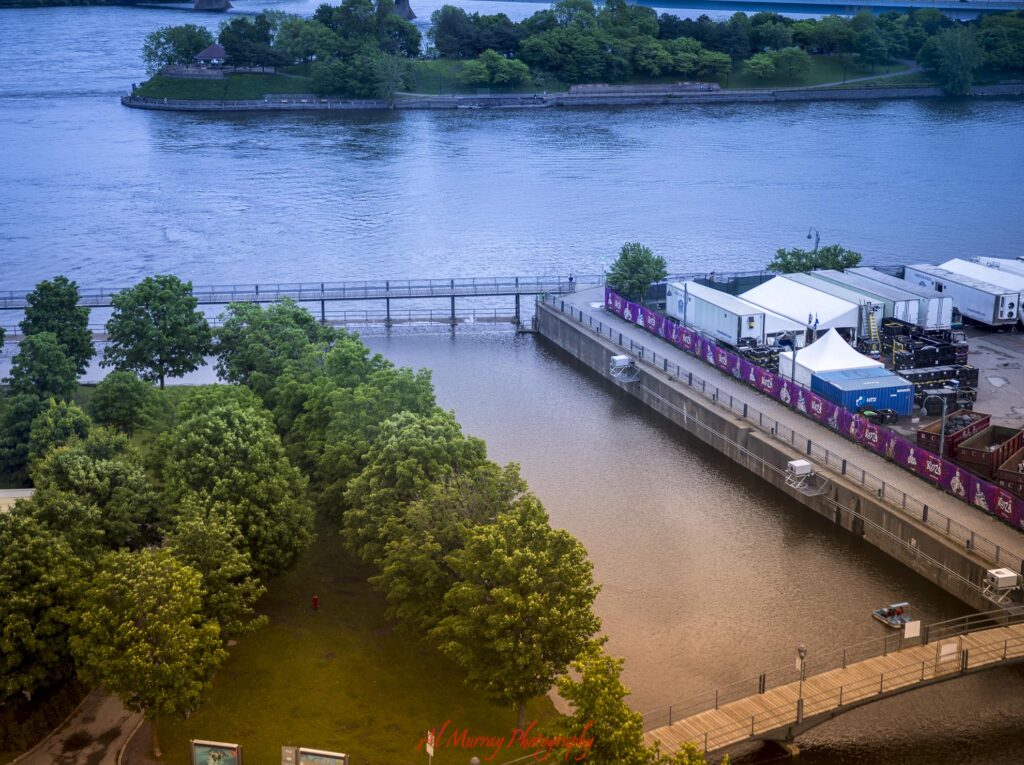 Elevated view over the Montreal waterfront with a tree filled park. pedestrian paths. event structures. and the wide river beyond. The waterfront scenery offers useful context when deciding should I stay in Old Montreal or Downtown.