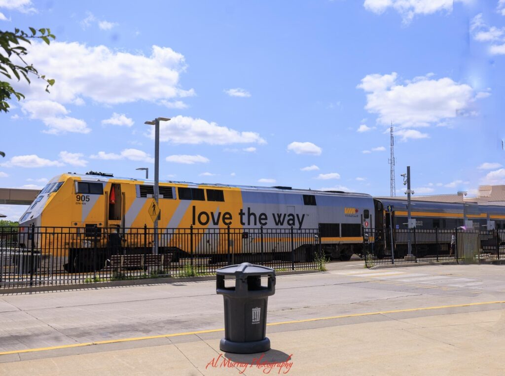 A yellow and silver VIA Rail train with the words "love the way" waits beside a station platform under a bright blue sky. The train represents rail transportation for a day trip to Guelph from Toronto.