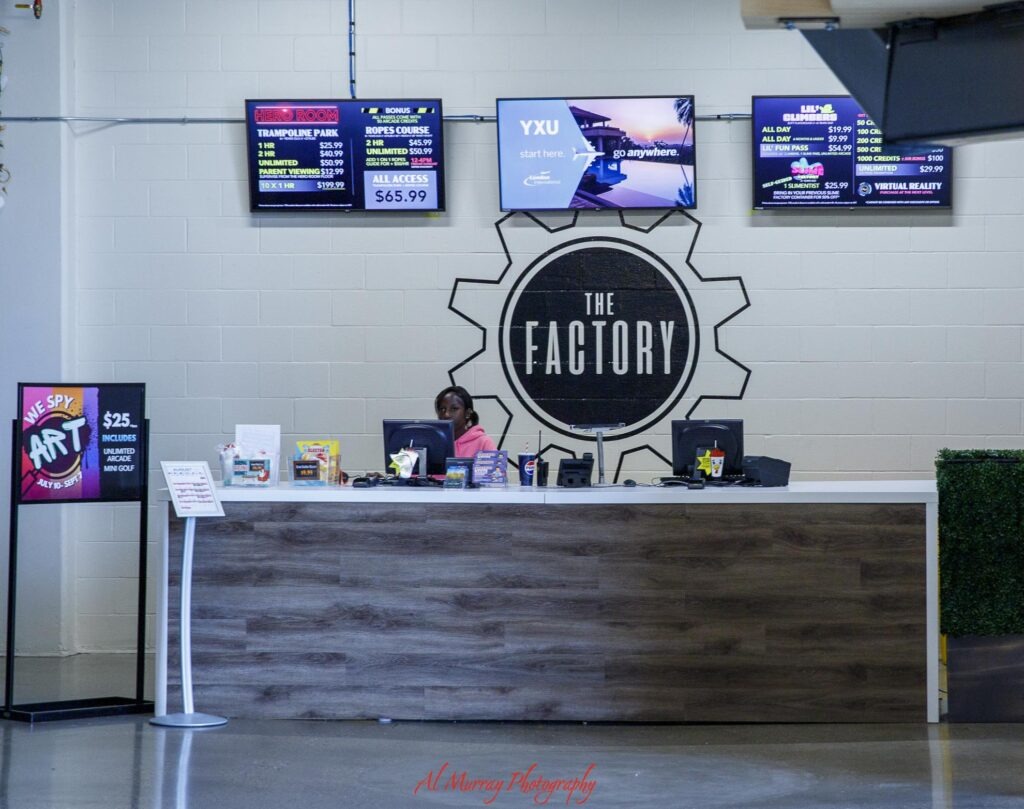 The front desk at The Factory in London Ontario sits beneath a large gear shaped logo reading "THE FACTORY" with digital screens advertising activities and prices. Readable signs include "HERO ROOM". "TRAMPOLINE PARK". "1 HR $25.99". "2 HR $40.99". "UNLIMITED $50.99". "PARENT VIEWING $12.99". "10 X 1 HR $199.99". "ROPES COURSE". "2 HR $45.99". "ALL ACCESS $65.99". "YXU". "start here". "go anywhere". "London International Airport". "LIL CLIMBERS". "ALL DAY $19.99". "LIL FUN PASS $54.99". "VIRTUAL REALITY". "WE SPY ART". "$25". "INCLUDES UNLIMITED ARCADE MINI GOLF". Smaller pricing text is present but not fully legible. The indoor attraction offers several activities that can be part of a London Ontario day trip.
