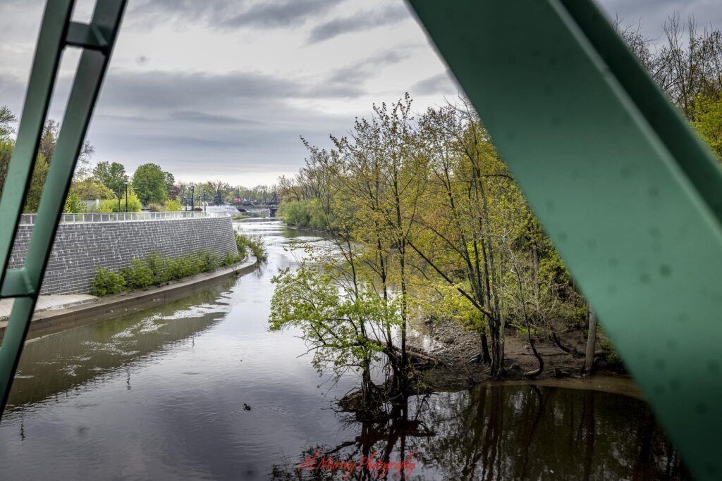 A calm river winds through a wooded area in London Ontario beneath an overcast sky with large green bridge beams framing the view. The riverside scenery highlights the quieter outdoor side of a London Ontario weekend getaway.