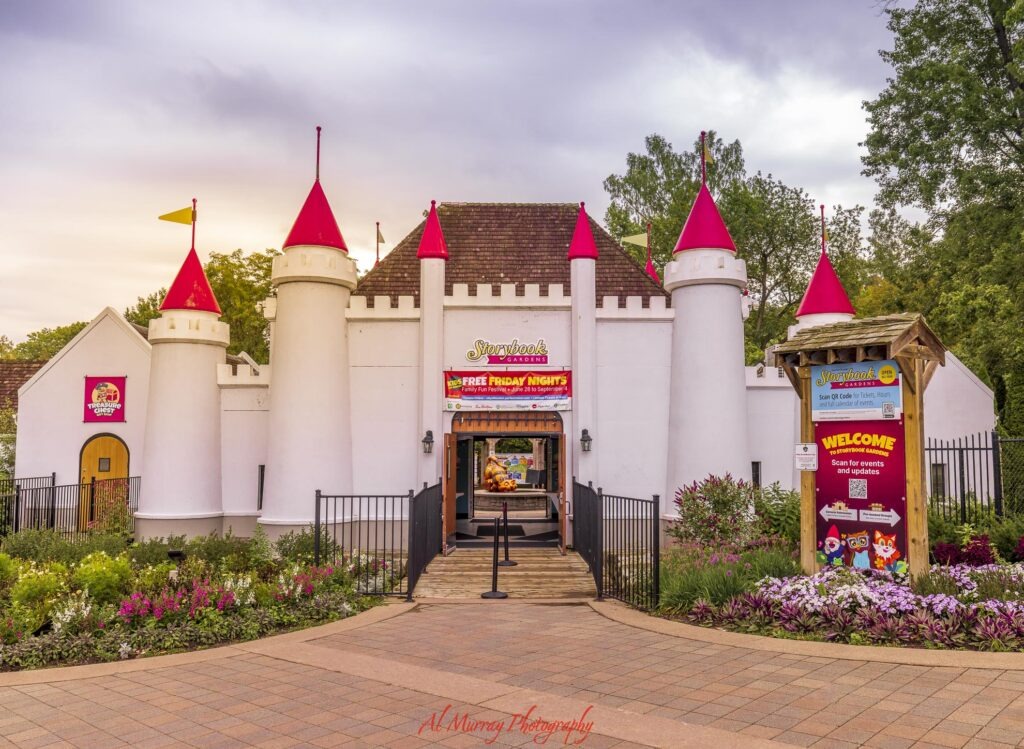 The white castle entrance to Storybook Gardens in London Ontario has bright red turret roofs and colorful flower beds surrounding the walkway. Signs read "Storybook Gardens". "Kids Free Friday Nights". "Family Fun Festival June 26 to September 4". "Treasure Chest Gift Shop". "Welcome to Storybook Gardens". "Scan for events and updates". This family attraction is a playful stop during a weekend in London Ontario.