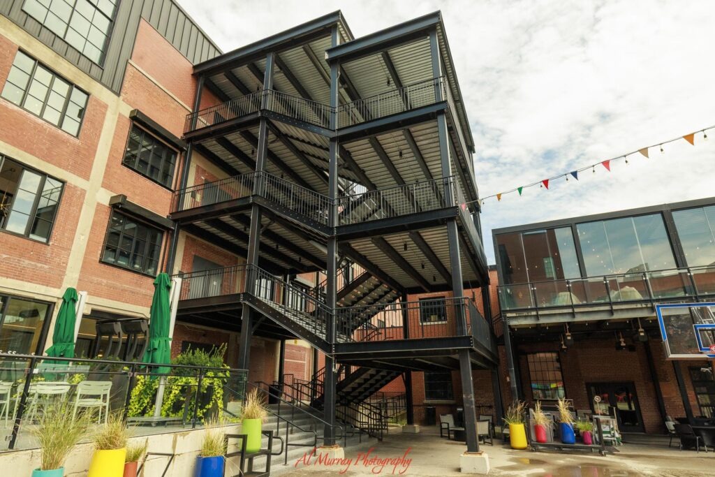 A renovated red brick industrial building in London Ontario features black steel staircases balconies patio seating and colorful planters in an open courtyard. The mix of historic architecture and modern gathering spaces adds to the appeal for travelers wondering if London Ontario is worth visiting